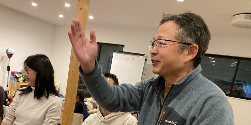 夜の食卓で、男性が手を挙げて横の人に話している写真